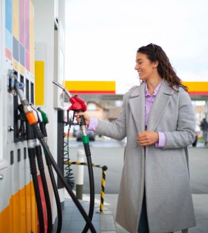Woman in a gray coat fueling a car at a gas station, smiling while holding a red fuel nozzle to the vehicle's tank.