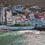 Colorful hillside coastal town with multicolored apartment buildings above a turquoise sea and rocky hills in the background.