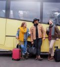 772656 - Grupo Dis Servicios | Upitravel Three travelers with rolling suitcases chat and smile beside a yellow bus outside a street.