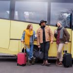 772656 - Grupo Dis Servicios | Upitravel Three travelers with rolling suitcases chat and smile beside a yellow bus outside a street.
