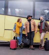 772656 - Grupo Dis Servicios | Upitravel Three travelers with rolling suitcases chat and smile beside a yellow bus outside a street.