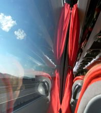 Viewing countryside while traveling with bus | Upitravel Bus interior with red curtains and red seats, window showing highway and hills outside.