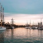 Barcelona port at twilight | Upitravel Marina at sunset with sailboats docked along both sides and palm trees lining the shore.