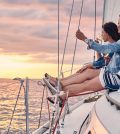 Two friends sit on a sailboat's bow at sunset, each holding a glass of wine and enjoying the view over calm waters.