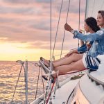 Two friends sit on a sailboat's bow at sunset, each holding a glass of wine and enjoying the view over calm waters.