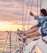 Two friends sit on a sailboat's bow at sunset, each holding a glass of wine and enjoying the view over calm waters.
