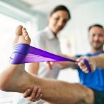physiotherapist doing treatment with patient in bright office | Upitravel Therapist assists a patient in a leg stretch using a purple resistance band.