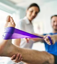 physiotherapist doing treatment with patient in bright office | Upitravel Therapist assists a patient in a leg stretch using a purple resistance band.