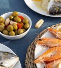 778927 - Restaurante Las Palmas | Upitravel Plate of orange-pasted shrimp with lemon wedges on a woven mat; a bowl of green olives and a whole fish on another plate visible nearby.