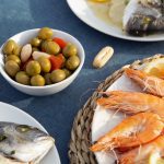 778927 - Restaurante Las Palmas | Upitravel Plate of orange-pasted shrimp with lemon wedges on a woven mat; a bowl of green olives and a whole fish on another plate visible nearby.