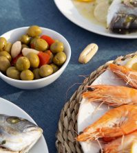 778927 - Restaurante Las Palmas | Upitravel Plate of orange-pasted shrimp with lemon wedges on a woven mat; a bowl of green olives and a whole fish on another plate visible nearby.