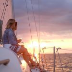 785141 - Kaisailing | Upitravel Two people sit on a sailboat at sunset, holding drinks and watching the horizon.
