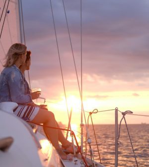 785141 - Kaisailing | Upitravel Two people sit on a sailboat at sunset, holding drinks and watching the horizon.